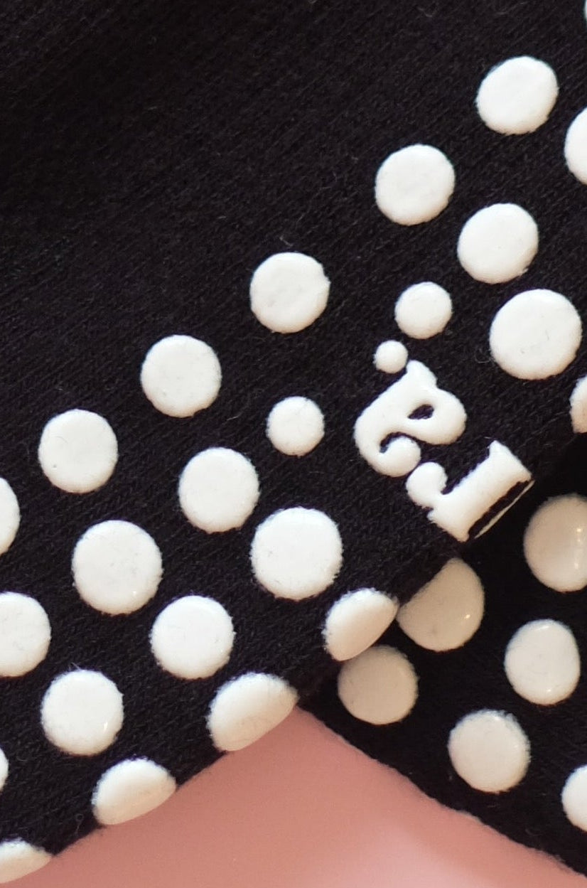 A pair of black grip socks with white polka dots on the toes and heels, laid flat on a solid pink background. The brand name 'nut & studio' is printed in white on the side of each sock.